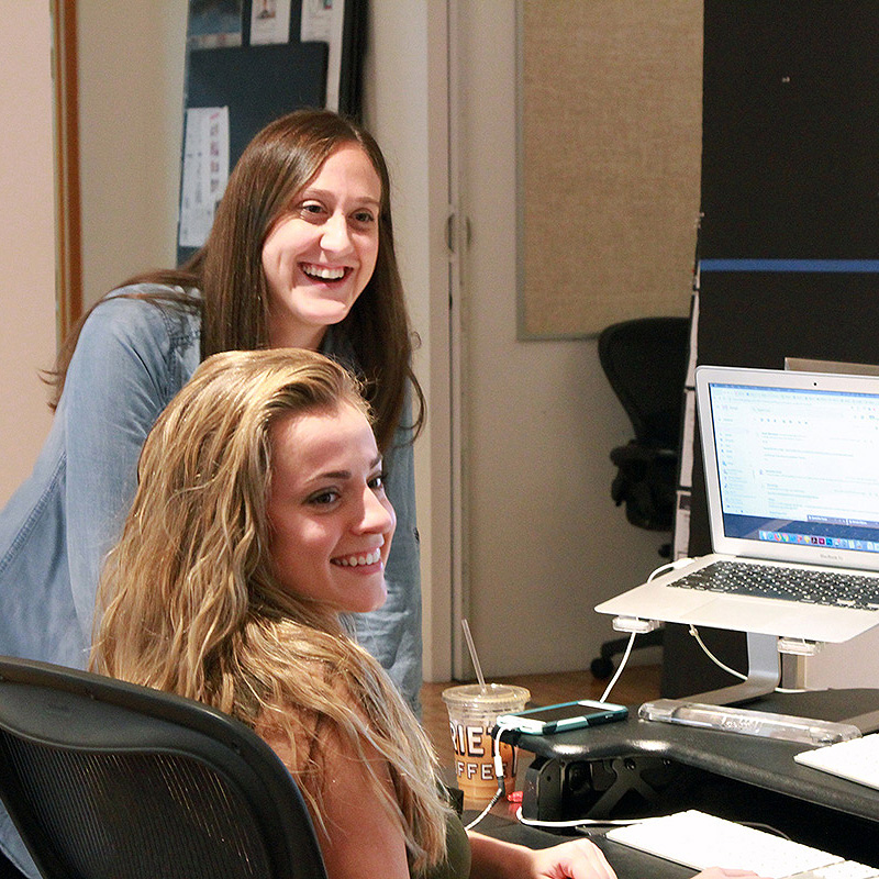 Two women smile while sitting at and standing near a desk with a laptop.
