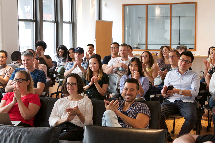 A large group of diverse people smile, clap and cheer while sitting at a company meeting.